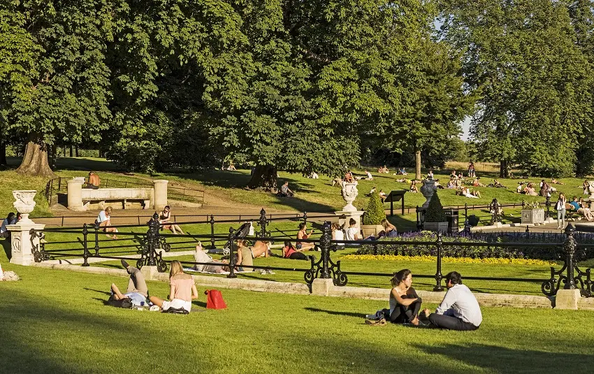 Människor som sitter utspridda på gräset och på bänkar i en solig Kensington Park